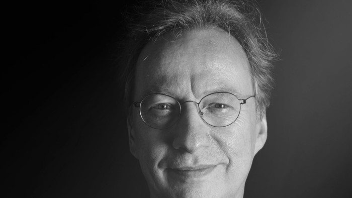 Ein älterer Mann mit Brille und dunkler Kleidung lächelt in die Kamera.