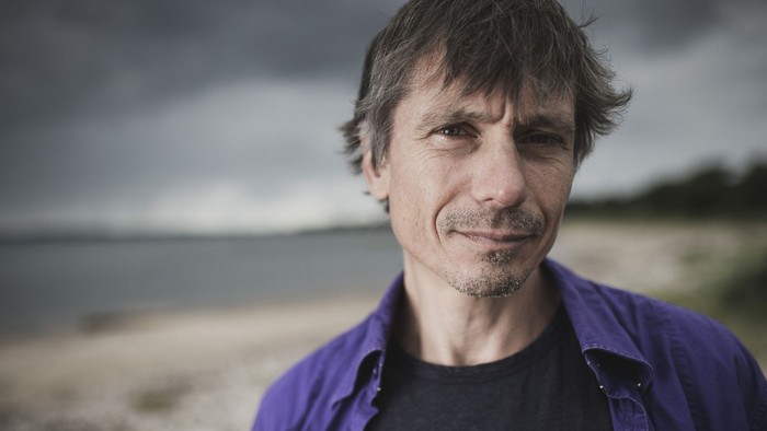 En mand med et eftertænksomt udtryk står ved en strand under en dramatisk, skyet himmel.