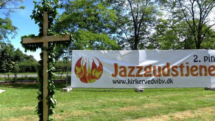 En grøn plads med en skiltet med Jazzgudstienens logo og website, og et kryds med planter.