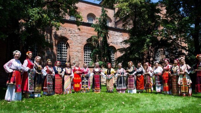 En gruppe kvinder i traditionelle kostumer står i en kreds på en eng og synger.