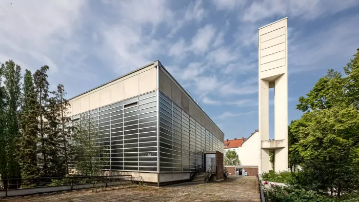 Moderner Gebäudekomplex mit Glasfassade und hohem weißem Turm, umgeben von Grünflächen.