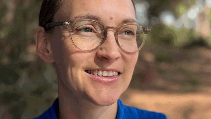 En smilende person med briller og en blå bluse udendørs i naturligt dagslys.