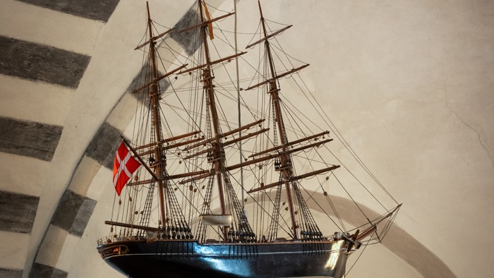 Et modelleskib hænger fra tak i en væg med bueformede vinduer og flag.