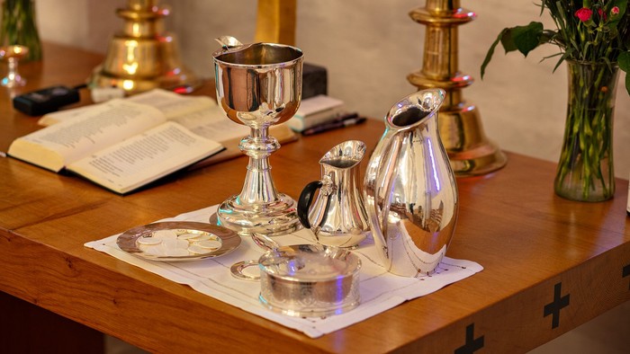 Hellig sølvtøj og kar arrangeret på et træbord med religiøse genstande og blomster.