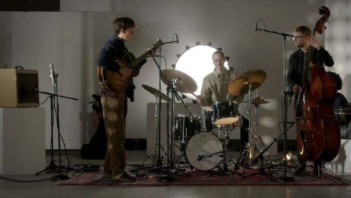 Et trio af musikere optræder live med elguitar, trommer og kontrabas i et svagt oplyst lokale.