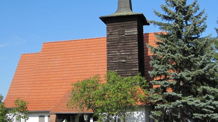 Kirchengebäude mit hohem Turm und rotem Dach, umgeben von Bäumen und Büschen