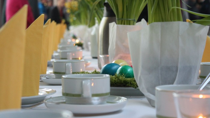 Ein festlich gedeckter Tisch mit weißen Tellern, Tassen und grünen Ostereiern, begleitet von gelben Servietten und Topfpflanzen