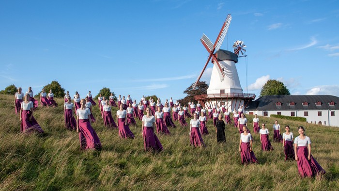 En stor gruppe i matchende bordeauxrøde kjoler udfører koreografi nær en traditionel vindmølle på en græsklædt bakkeside.