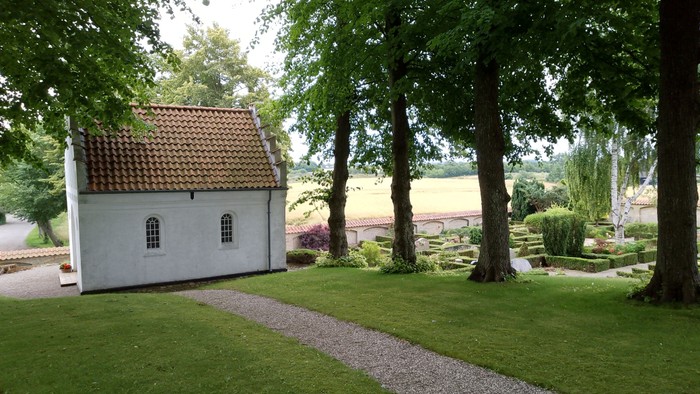 Lille hvid bygning med rødt tag, omgivet af træer og græs.