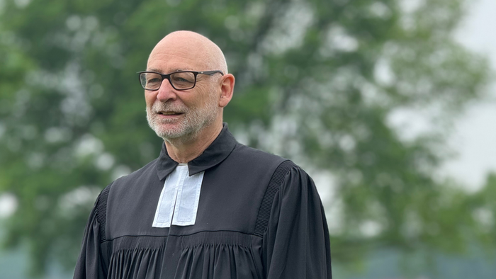 Ein glatzköpfiger Mann mit Brille trägt im Freien eine schwarze Richterrobe vor einem unscharfen grünen Waldhintergrund.