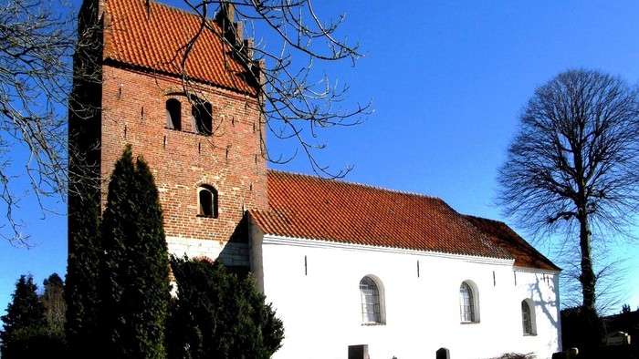 En hvid kirke med røde tag og tårn, omgivet af træer og buske under et klarblåt himmel.