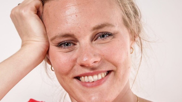 En glad kvinde med blondt hår, der smiler, mens hun hviler hånden på hovedet i en rød top.