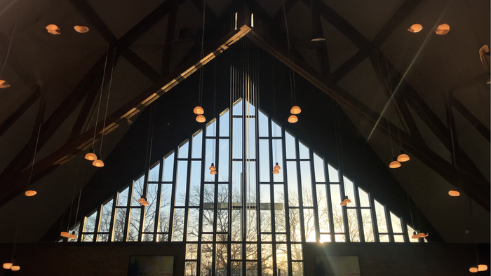 Moderne kirkeinteriør med høje trekantede glasmosaikvinduer og varme hængende lysarmaturer.