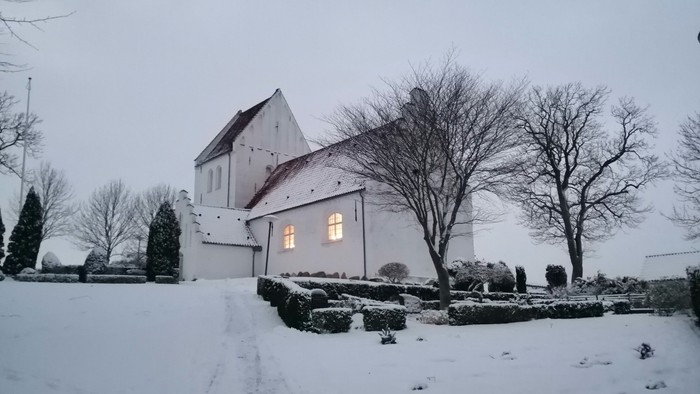 En snedækket kirke med et lysende vindue og nakke træer.