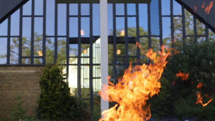 En levende ild brænder klart i et udendørs bålsted nær et stort kors og en bygning med glaspaneler.