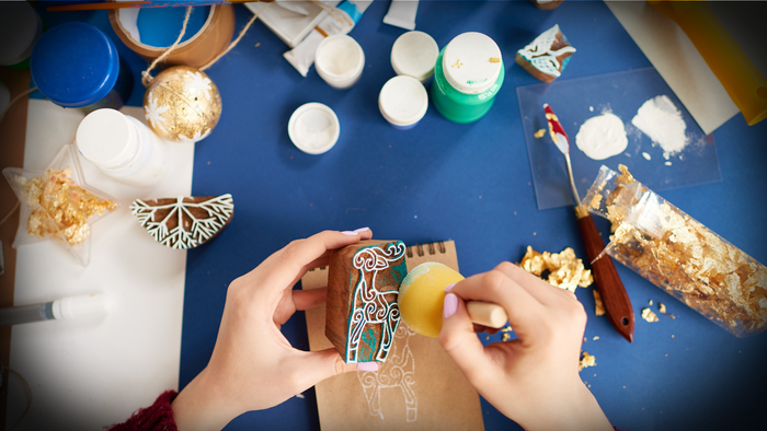 Zwei Hände basteln an einem weihnachtlich bemalten Holzpferd auf einem Tisch mit Pinseln, Farben, Goldfolie und Bastelmaterialien. Im Hintergrund liegen Weihnachtsdekorationen und Farbtöpfe – eine gemütliche kreative Adventsszene.