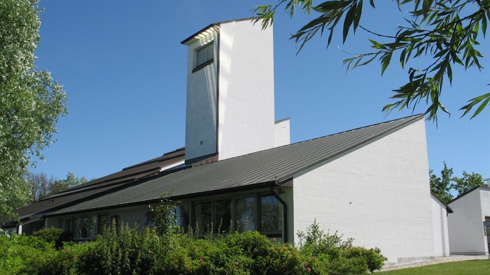 Moderne hvid bygning med store vinduer og et højt tårn, omgivet af grønne områder.