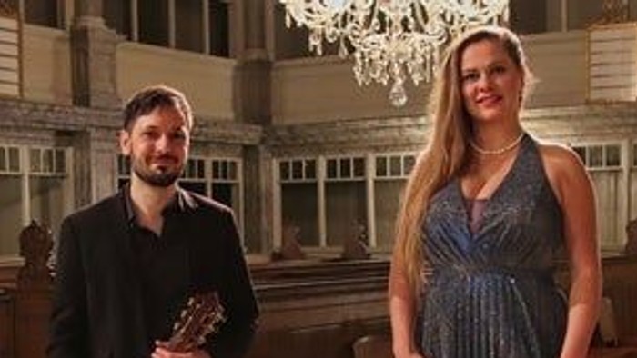 To musikere poser sammen, den ene holder en guitar og den anden er iført elegant tøj, i en prægtig indendørs omgivelse.