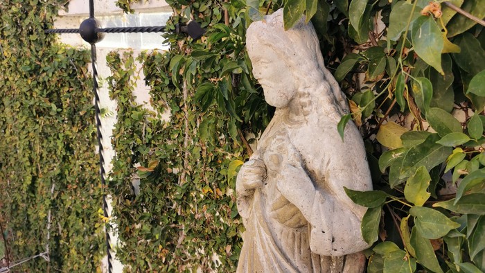 Die Statue einer Frau mit einem Vogel auf dem Arm, umgeben von grünem Blattwerk.