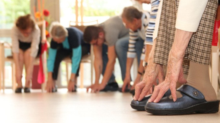 Eine Gruppe älterer Menschen nimmt an einem Fitnesskurs teil, alle beugen sich nach vorne.