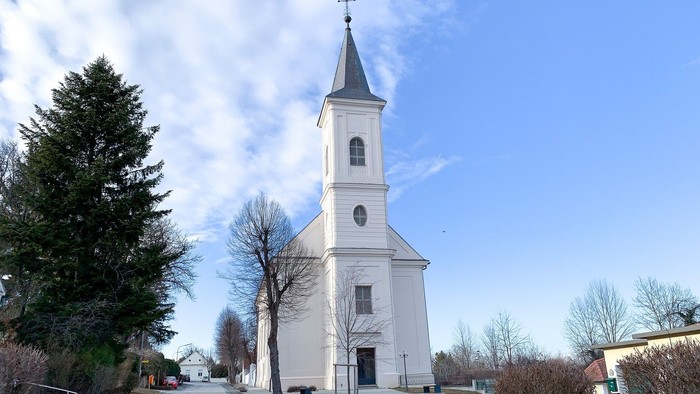 Weiße Kirche mit hohem Turm und Uhr, umgeben von Bäumen und Himmel.