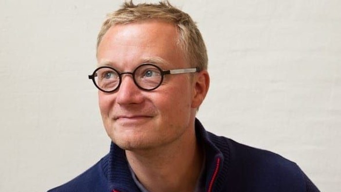 En mand med kort, lyst hår, iført runde briller og en mørkeblå sweater med rød kant, der smiler let.