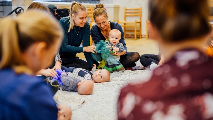 En gruppe kvinder sidder på gulvmatte og omhandler børnepuppetter.