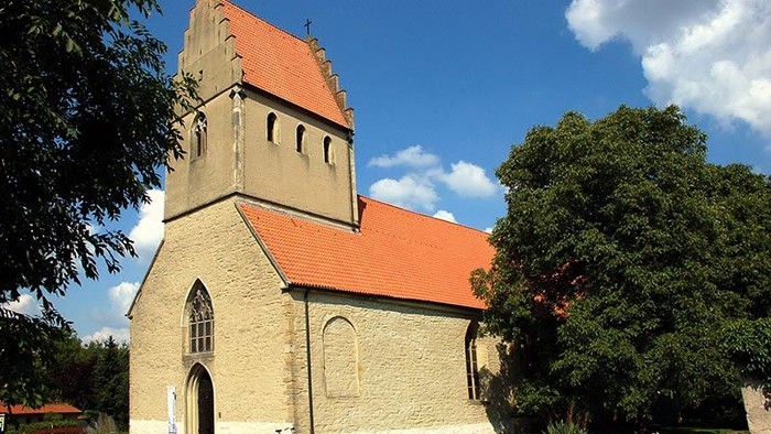 Kirchengebäude mit hohem Turm und rotem Dach, umgeben von Bäumen und Blumenbeeten
