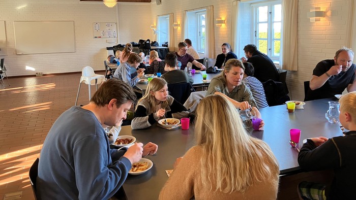 En gruppe mennesker, der spiser ved lange borde i et veloplyst rum.