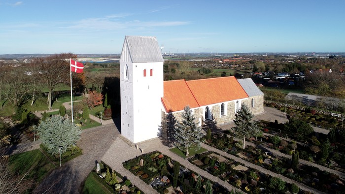 En lille kirke med tårn og rødt tag, beliggende i en landsby med en dansk flag.