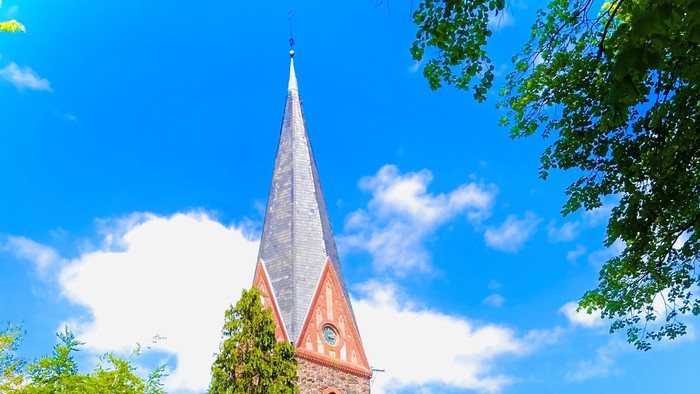Ein hohes, spitzes Kirchenturm aus rotem Backstein, umgeben von üppigem Grün und einem klaren blauen Himmel