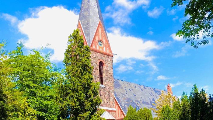 Ein hohes, spitzes Kirchenturm aus rotem Backstein, umgeben von üppigem Grün und einem klaren blauen Himmel
