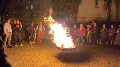Die Gruppe versammelt sich um ein großes Lagerfeuer in einer städtischen Freiluftumgebung bei Nacht.