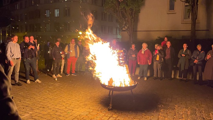 Die Gruppe versammelt sich um ein großes Lagerfeuer in einer städtischen Freiluftumgebung bei Nacht.