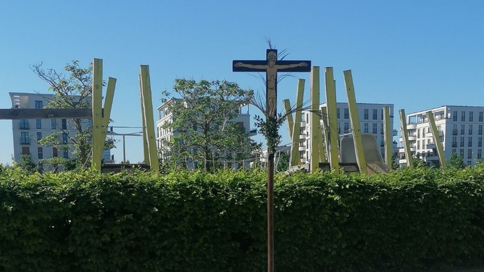 Ein hölzernes Kreuz steht auf einem Betonpfahl im Freien, umgeben von Büschen und Gebäuden im Hintergrund.