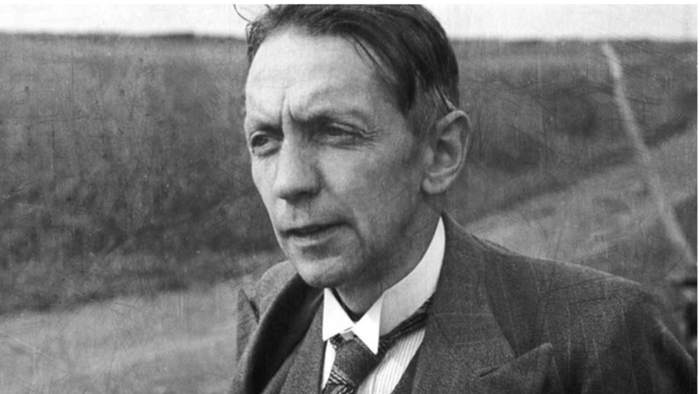 En alvorligt udseende mand i jakkesæt og slips poser udendørs på et gammelt sort-hvidt fotografi.