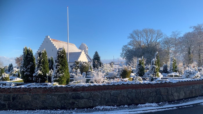 En snedækket kirkegård med en kirke i baggrunden og en mur foran.
