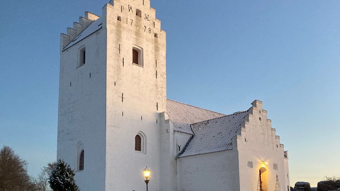 En snede hvid kirke med tårn og en lystet indgang i snedæknede omgivelser