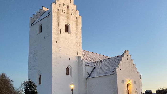 En snede hvid kirke med tårn og en lystet indgang i snedæknede omgivelser