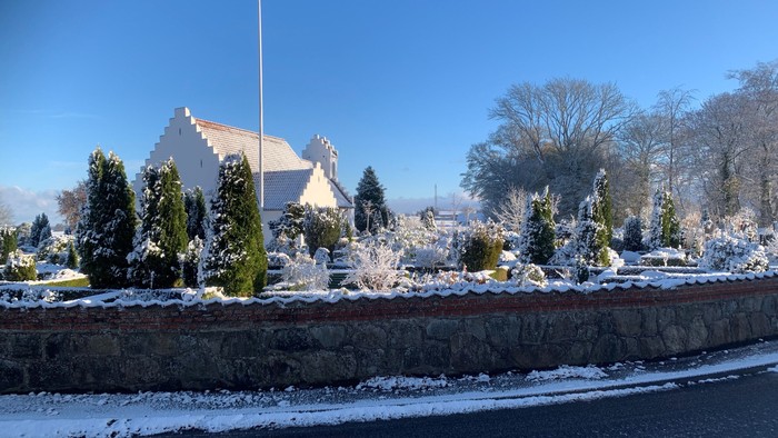En snedækket kirkegård med en kirke i baggrunden og en mur foran.