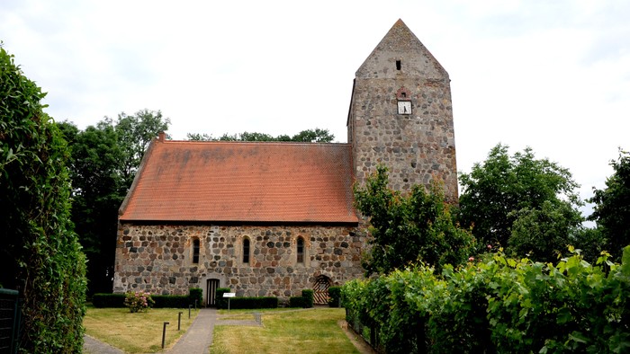 Alte Kirche mit hohem Turm und rotem Dach, umgeben von Bäumen und Büschen