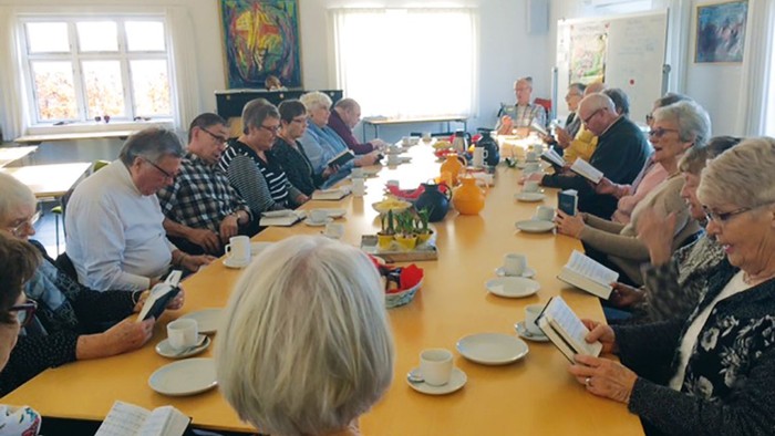 En gruppe mennesker sidder ved lange bord og spiser, mens de ser på mobiltelefoner.