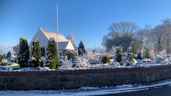 En snedækket kirkegård med en kirke i baggrunden og en mur foran.
