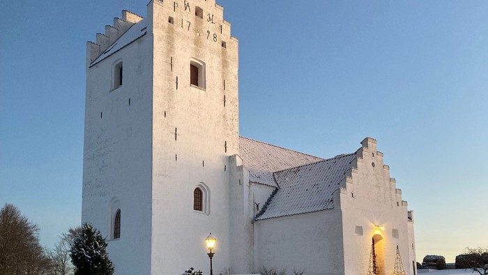 En snede hvid kirke med tårn og en lystet indgang i snedæknede omgivelser