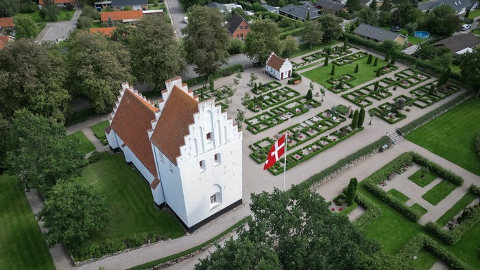 En hvid kastedal med røde tag og en dansk flag i have, omgivet af en formet have og boliger i baggrunden