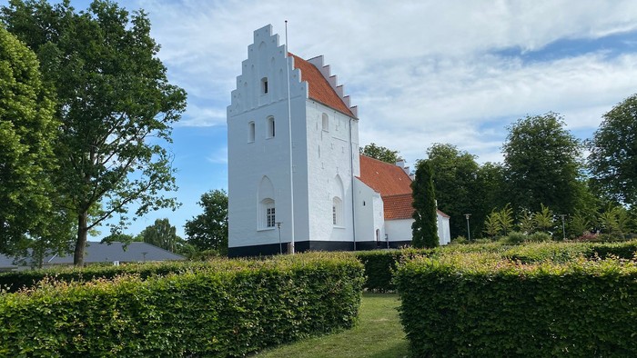 Et hvidt og orangenget tårn med hvide vinduer og røde tag, omgivet af grønne buske og træer under et blødt blå himmel.