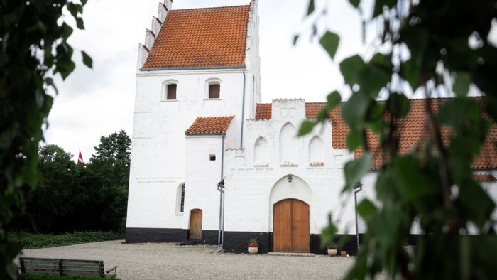 Et hvidt historisk tårn med røde tagfliser og trædør, omgivet af grønne buske og en benk.