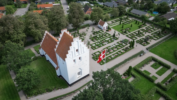 En hvid kastedal med røde tag og en dansk flag i have, omgivet af en formet have og boliger i baggrunden