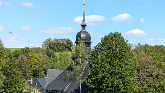 Kirchturm mit Kreuz auf grünem Hügel, umgeben von Bäumen und Häusern