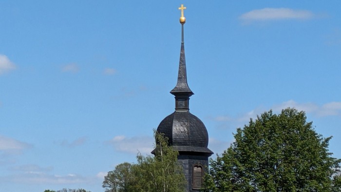 Ein schwarzer Kirchturm mit goldenem Kreuz vor blauem Himmel und Bäumen.